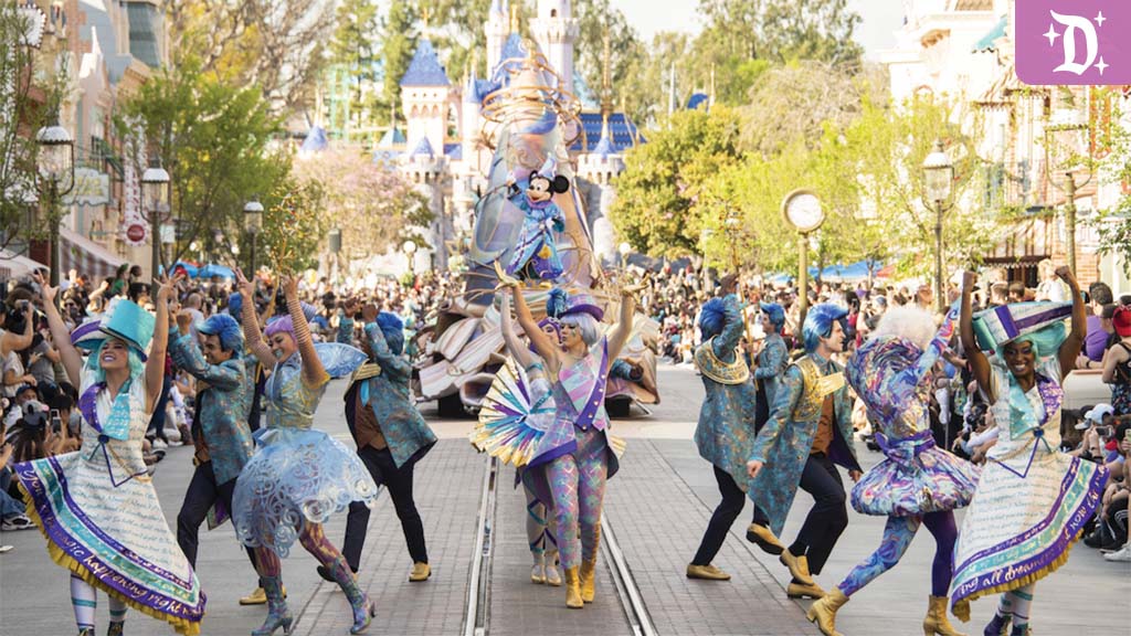 Dancers in iridescent costumes performing in a street parade with crowds on both sides, a float with a cartoon mouse figure and castle turrets behind.
