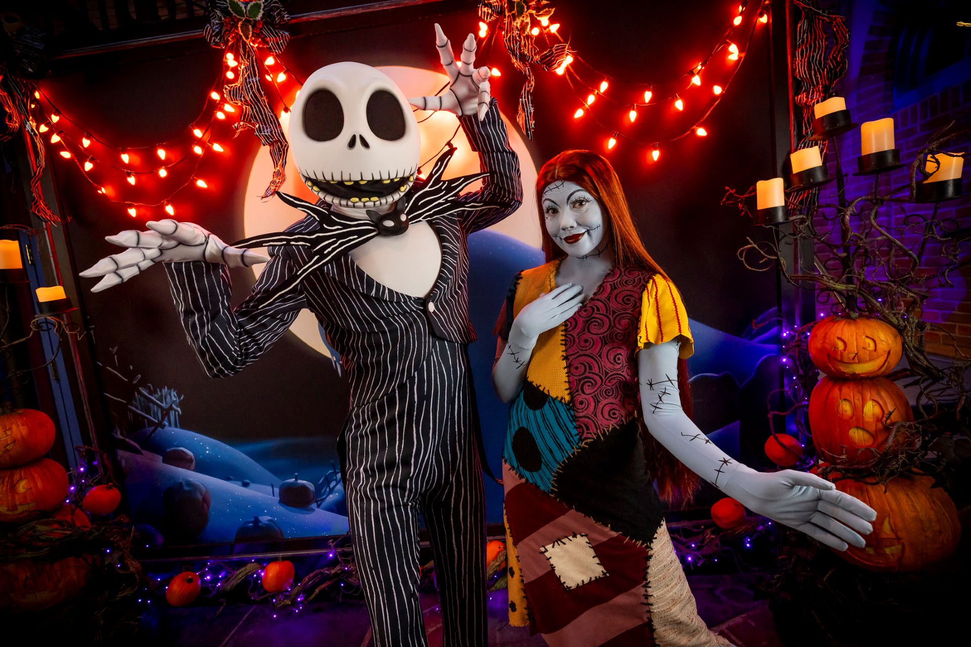 Skeleton-costumed figure in pinstriped suit and skull head beside a person in stitched makeup and patchwork dress among carved pumpkins and lights