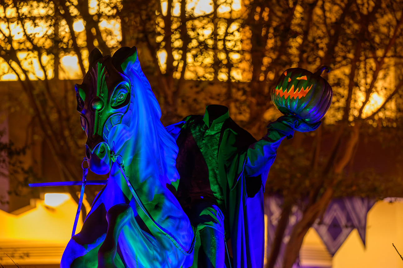 Headless horseman figure on a rearing horse holds a carved pumpkin under blue-green lights against a backdrop of trees