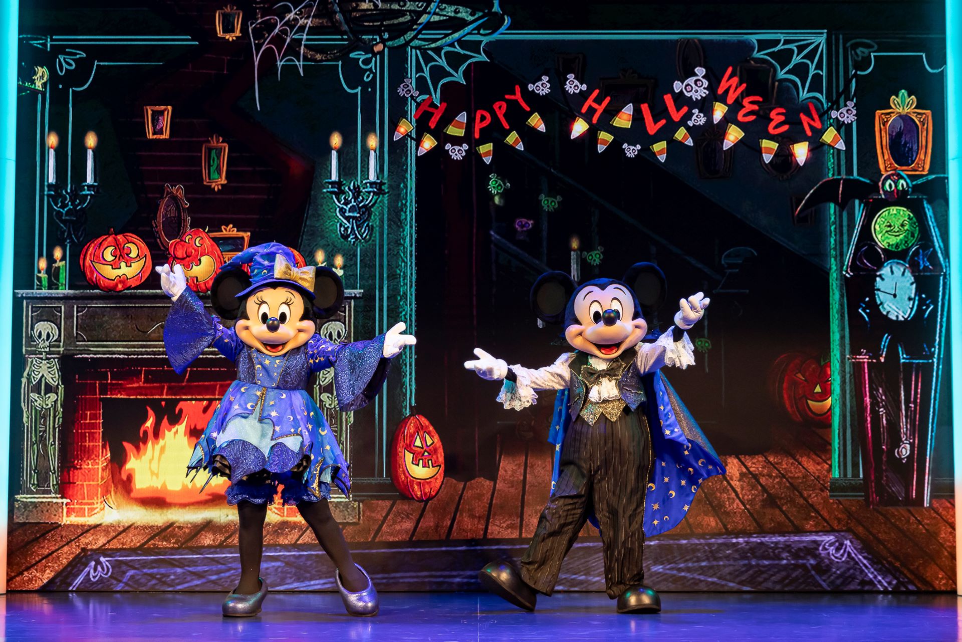 Two mouse mascots in blue wizard costumes on a stage with a lit fireplace, jack-o’lanterns, candles and a HAPPY HALLOWEEN banner