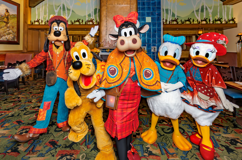 Goofy, Pluto, Clarabelle Cow, Donald Duck and Daisy Duck wearing autumn-themed outfits pose in a restaurant lobby with rustic décor