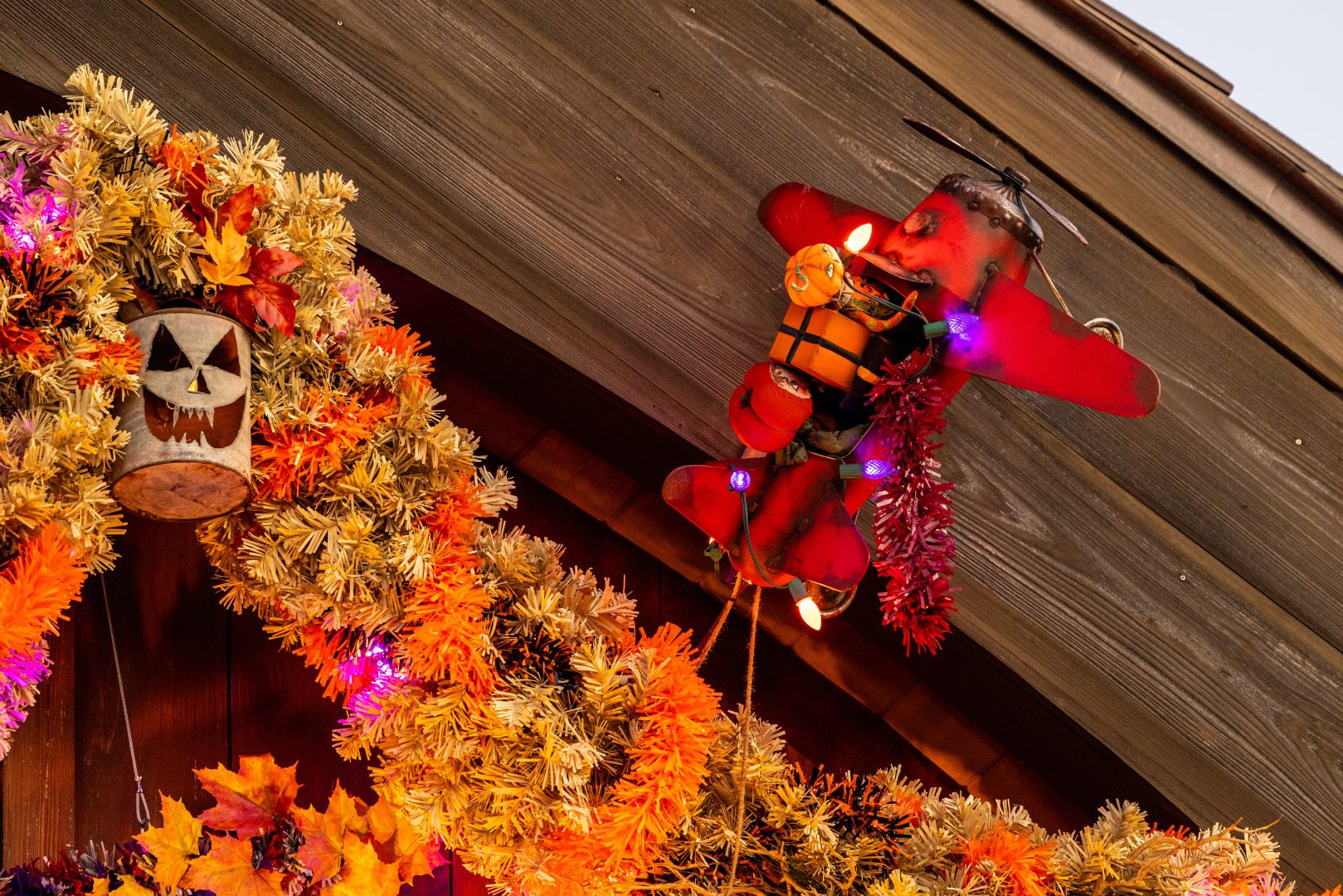 Rustic wooden eaves decorated with orange and gold tinsel garlands, purple lights, a metal jack-o'-lantern bucket, and a red toy airplane ornament