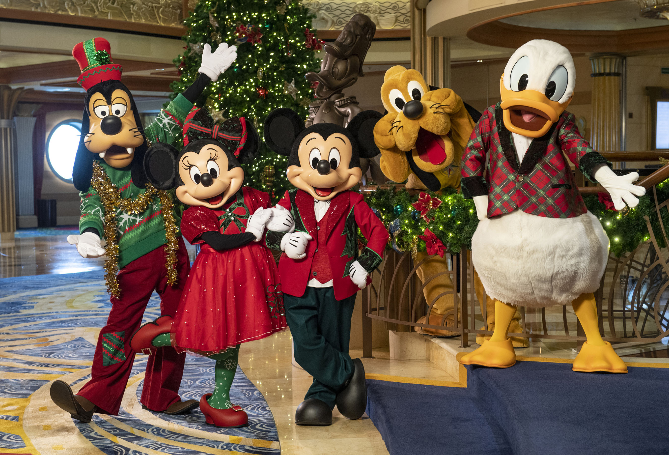 Disney characters Goofy, Minnie Mouse, Mickey Mouse, Pluto and Donald Duck in holiday outfits pose by a decorated Christmas tree on a cruise ship