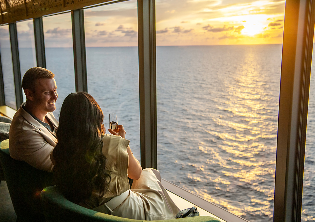 A man and woman seated by a large window on a ship at sunset, the woman holding a glass of champagne as they look out over the ocean.