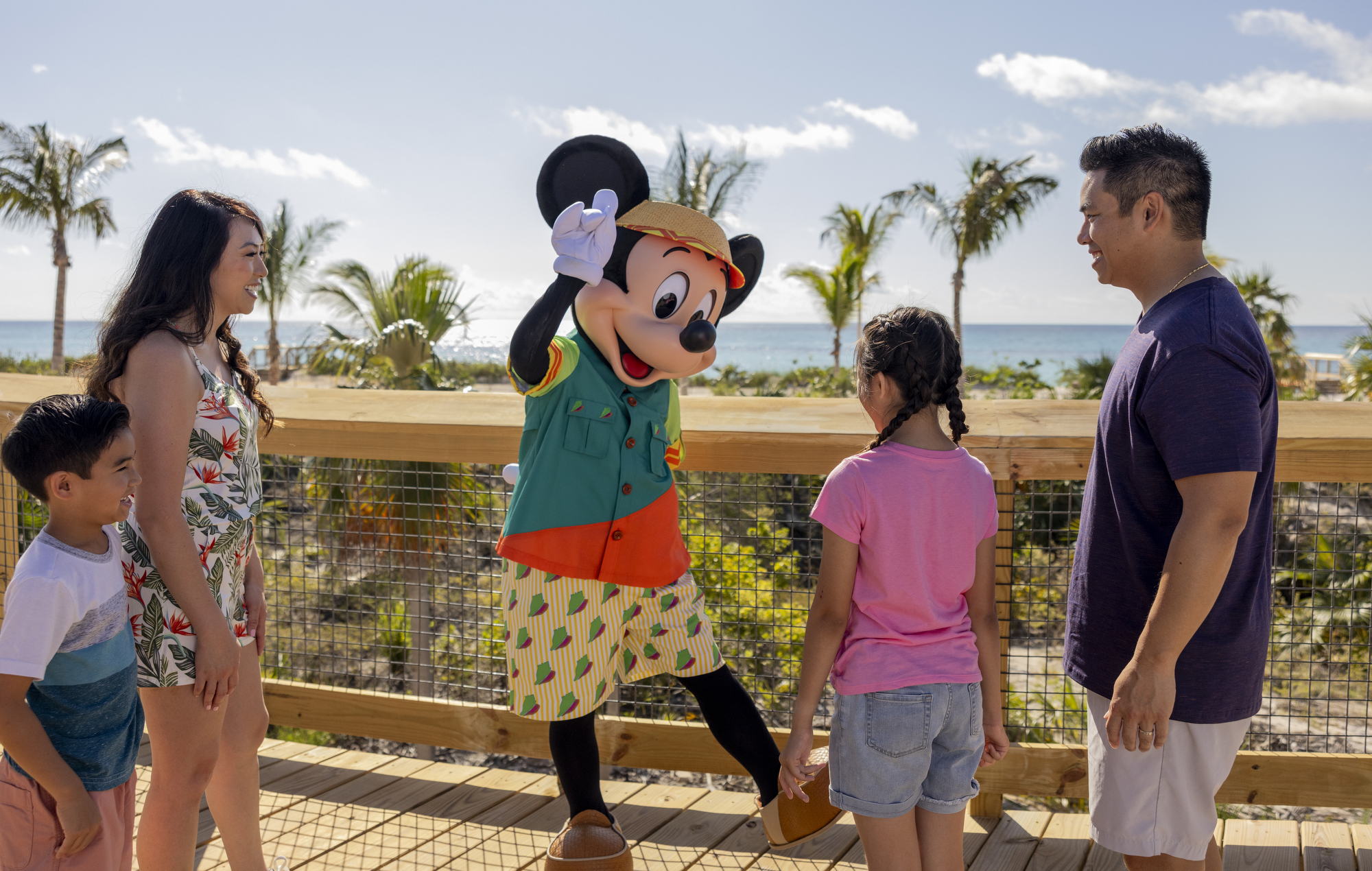 Family of four on a wooden boardwalk by the beach greeting Mickey Mouse in tropical outfit with palm trees and ocean in background.