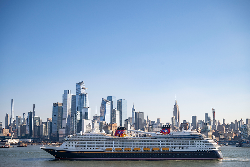 Cruise ship sailing past the Manhattan skyline of glass skyscrapers and a tall spire over a calm waterway under a clear blue sky