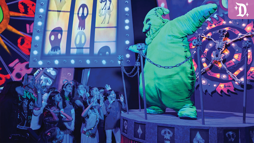 Group of children and adults watch an oversized green stitched-costume character on a raised stage with chains and a spinning skull wheel in dim light