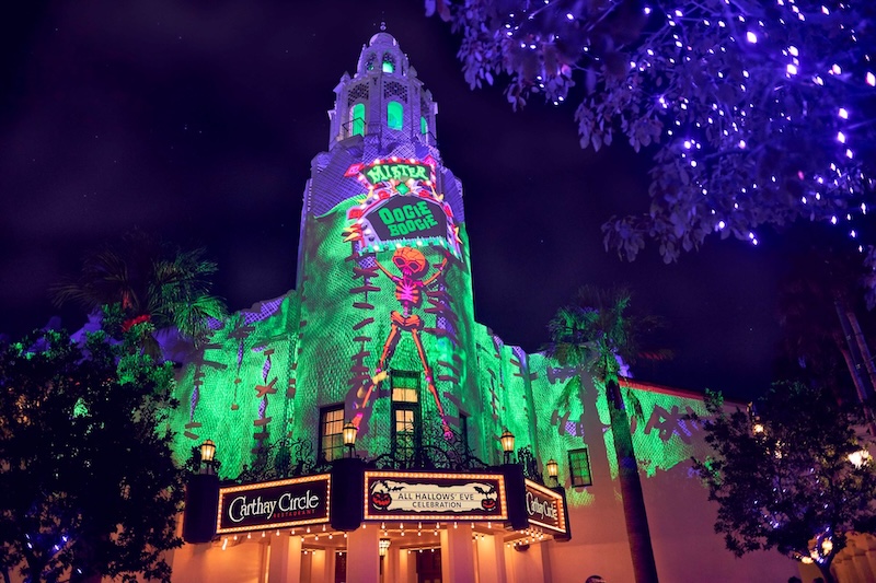 Tower lit green with a projected dancing skeleton above a Carthay Circle marquee, flanked by palm trees and branches lit in purple.
