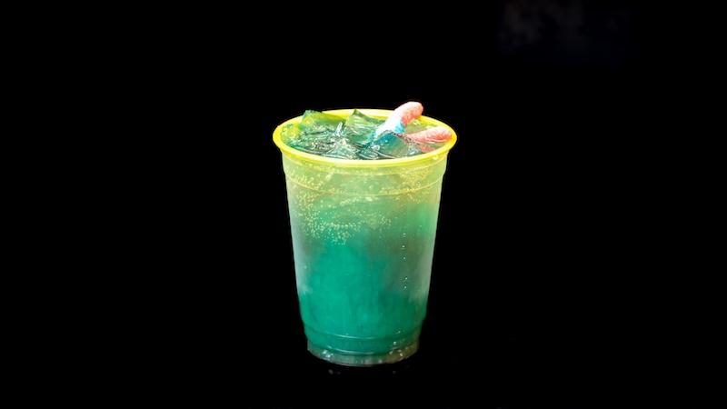 Translucent yellow plastic cup with greenish-blue fizzy drink, ice and red-and-blue gummy worms draped on the rim