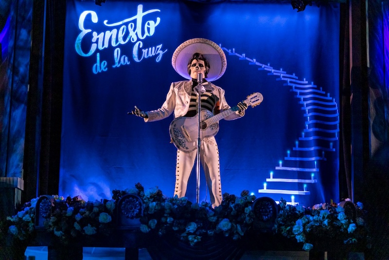 Skeleton mariachi in white suit and sombrero plays guitar at mic on flower-lined stage with neon staircase and Ernesto de la Cruz sign