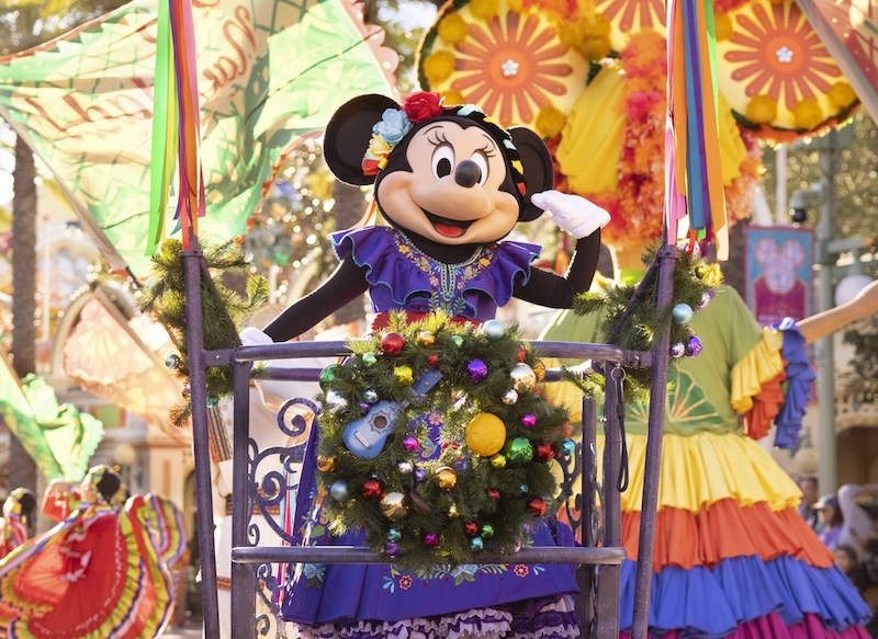 Minnie Mouse character in a purple dress and floral headpiece waving from a decorated parade float with wreath and ornaments.