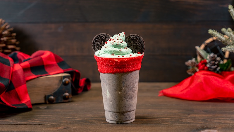 Cookies-and-cream milkshake with red sugar–coated rim, topped with green whipped cream and two Oreo cookies, on wooden table with holiday decor.