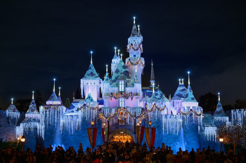 Fairytale-style castle illuminated in blue and white holiday lights with garlands and hanging icicle lights, nighttime crowd gathered below