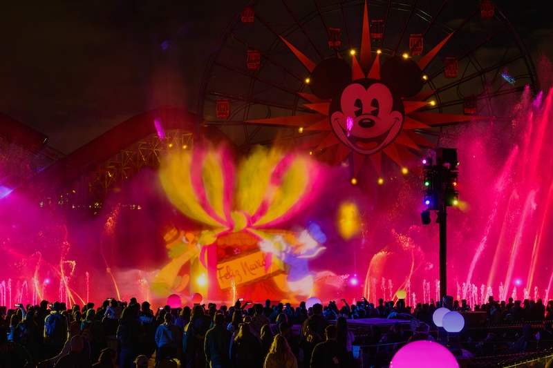 Crowd watches colorful nighttime water fountains and projections of a large gift with bow beneath a sun-shaped Ferris wheel lit in red and yellow