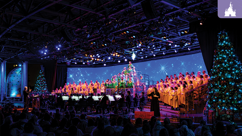 Choir wearing yellow and green robes sings on a lit stage with Christmas trees, backed by an orchestra under overhead stage lights