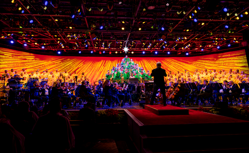 Conductor leading orchestra before a choir in green robes arranged in a pyramid shape under a glowing star on an orange-lit stage