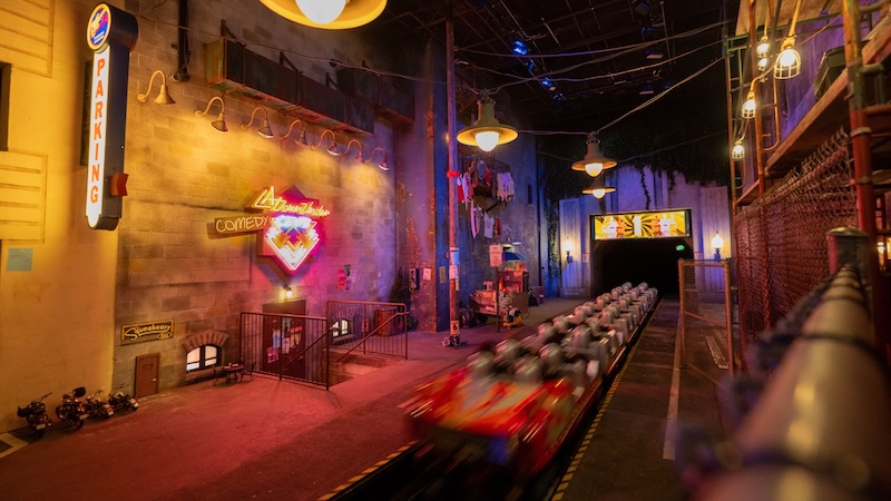 Blurred coaster train speeding through a neon-lit industrial station styled like an urban alley with overhead lamps and chain-link fencing