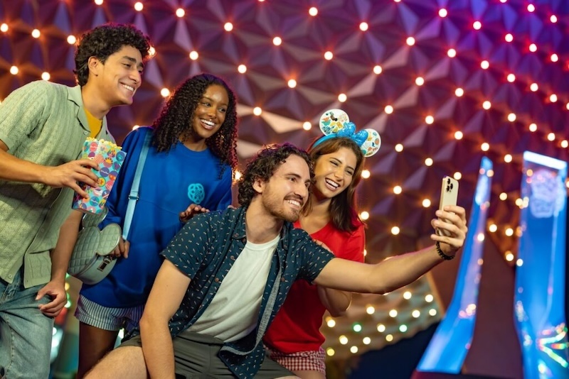 Four friends gathered under decorative lights, smiling as one extends a smartphone for a group selfie, one wearing a mouse-ear headband