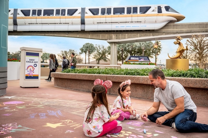 Father and two young girls in Minnie ears drawing hopscotch chalk art on pavement under a passing monorail at a theme park.