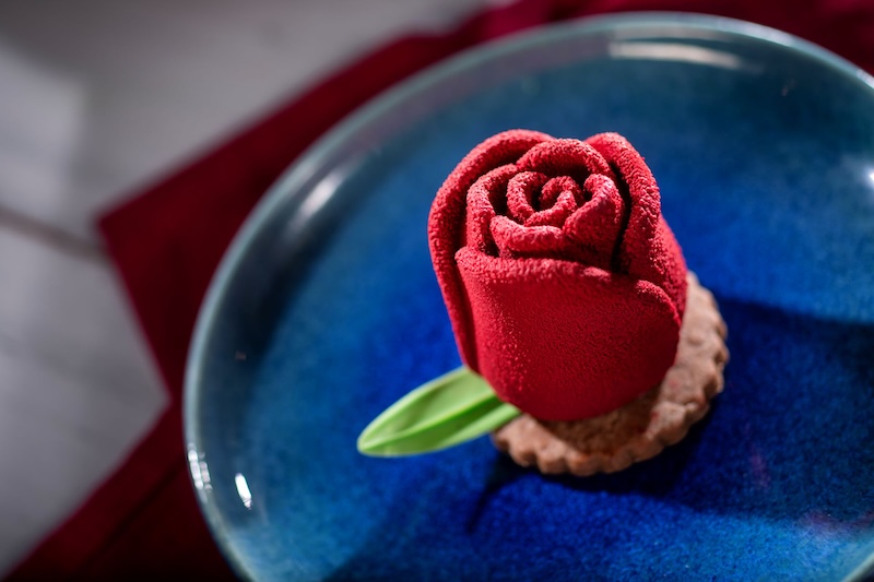 Red rose-shaped velvet-textured dessert on a crumbly cookie base with a green leaf garnish on a blue plate