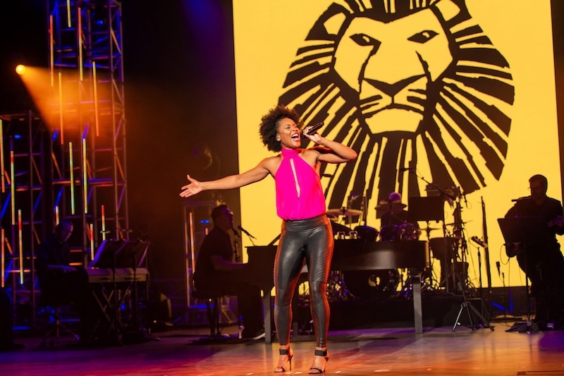Singer in pink top and black leather pants on stage with microphone in hand, large yellow screen with a stylized black lion head and band behind