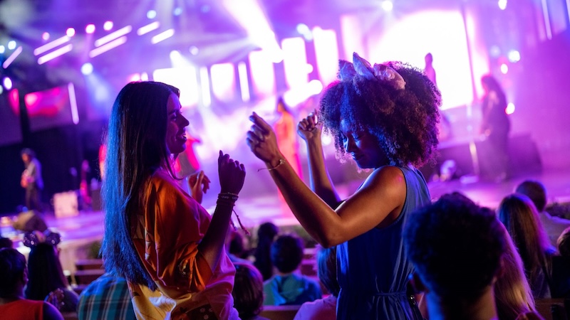 Two young women dancing in their seats, one wearing a cat-ear headband, under purple stage lights in a crowded music venue