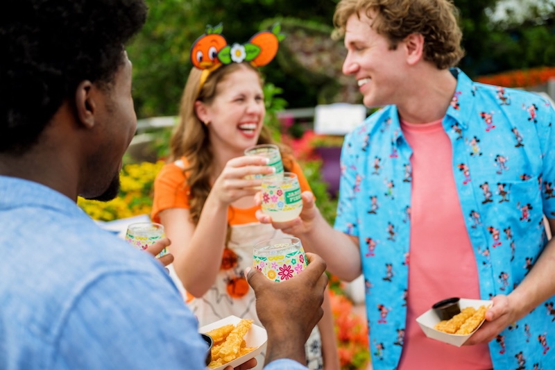 Three people in colorful clothes outdoors near flowers, raising decorative glasses of pale yellow beverage and holding trays of fried snacks