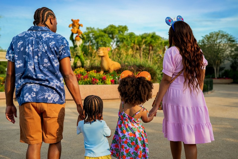 Family of four wearing mouse-ear headbands stands holding hands facing flowering animal topiaries in a garden plaza