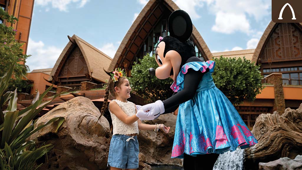 Young girl in denim shorts and flower headband smiling while holding hands with Minnie Mouse in a blue dress at a tropical resort courtyard.