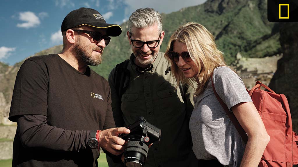Three travelers wearing sunglasses examine images on a DSLR camera against a backdrop of mountain ruins