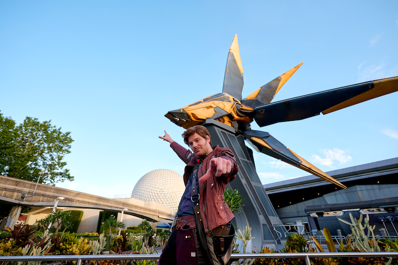 Man in red leather jacket pointing at camera in front of a white geodesic sphere and tall yellow-and-gray star-shaped structure