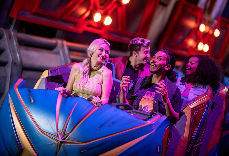 Four young adults seated in a blue ride vehicle with colorful lights overhead, smiling and raising their hands as the ride moves