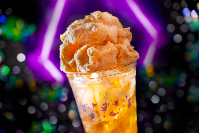 Clear plastic cup with orange iced drink and large mound of orange cotton candy on top, purple neon hexagon light and bokeh in background