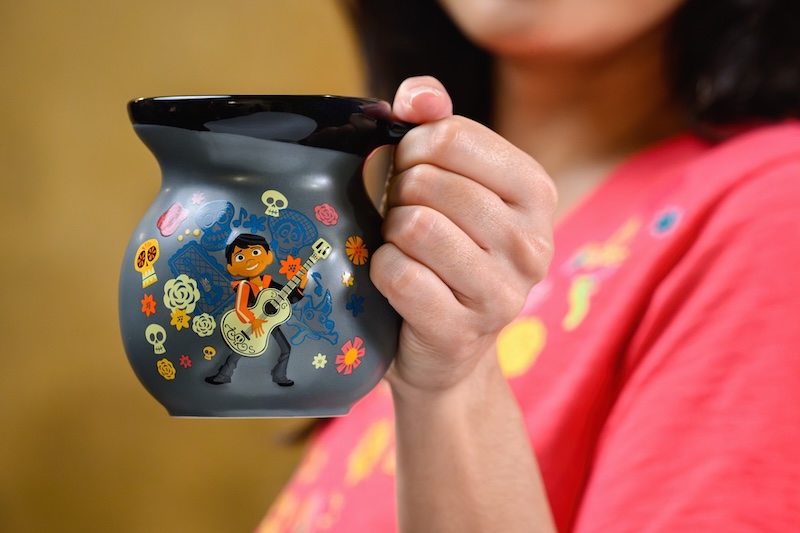 Hand holding a black ceramic mug decorated with a cartoon musician playing guitar surrounded by flowers and skulls