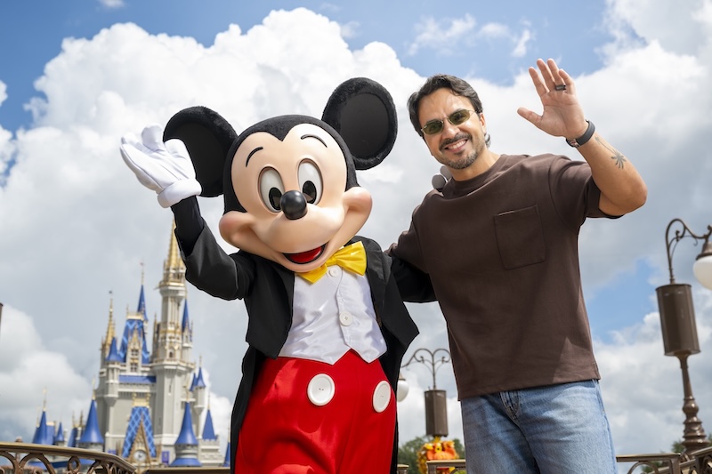 Man wearing brown t-shirt and sunglasses stands next to Mickey Mouse character in front of a castle, both waving at camera.