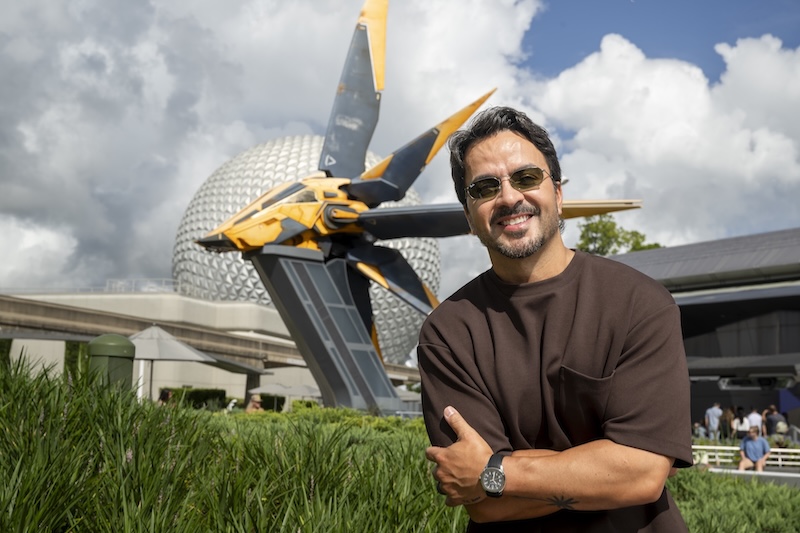Smiling man in sunglasses and brown shirt stands with arms crossed in front of a yellow sci-fi ship sculpture and geodesic dome at a park