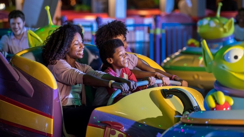 Mother and two children laugh and hold the safety bar as their glowing, alien-themed pods spin on an amusement ride.