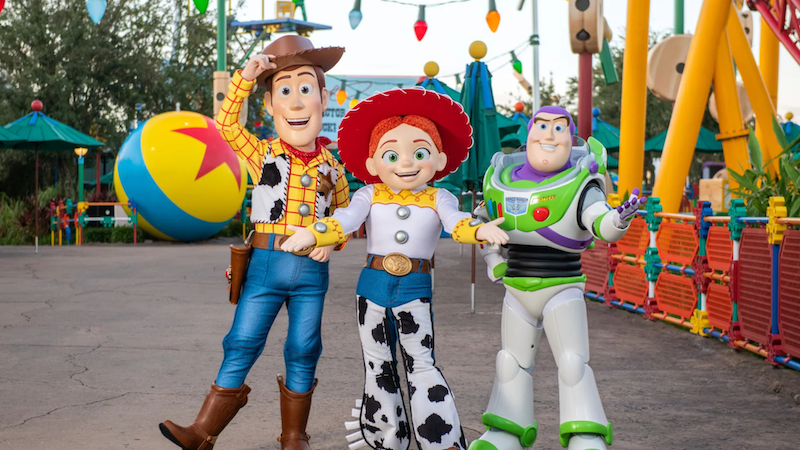 Three costumed characters – a cowboy, a cowgirl and an astronaut – stand arm in arm in a colorful outdoor theme park area with a giant toy ball
