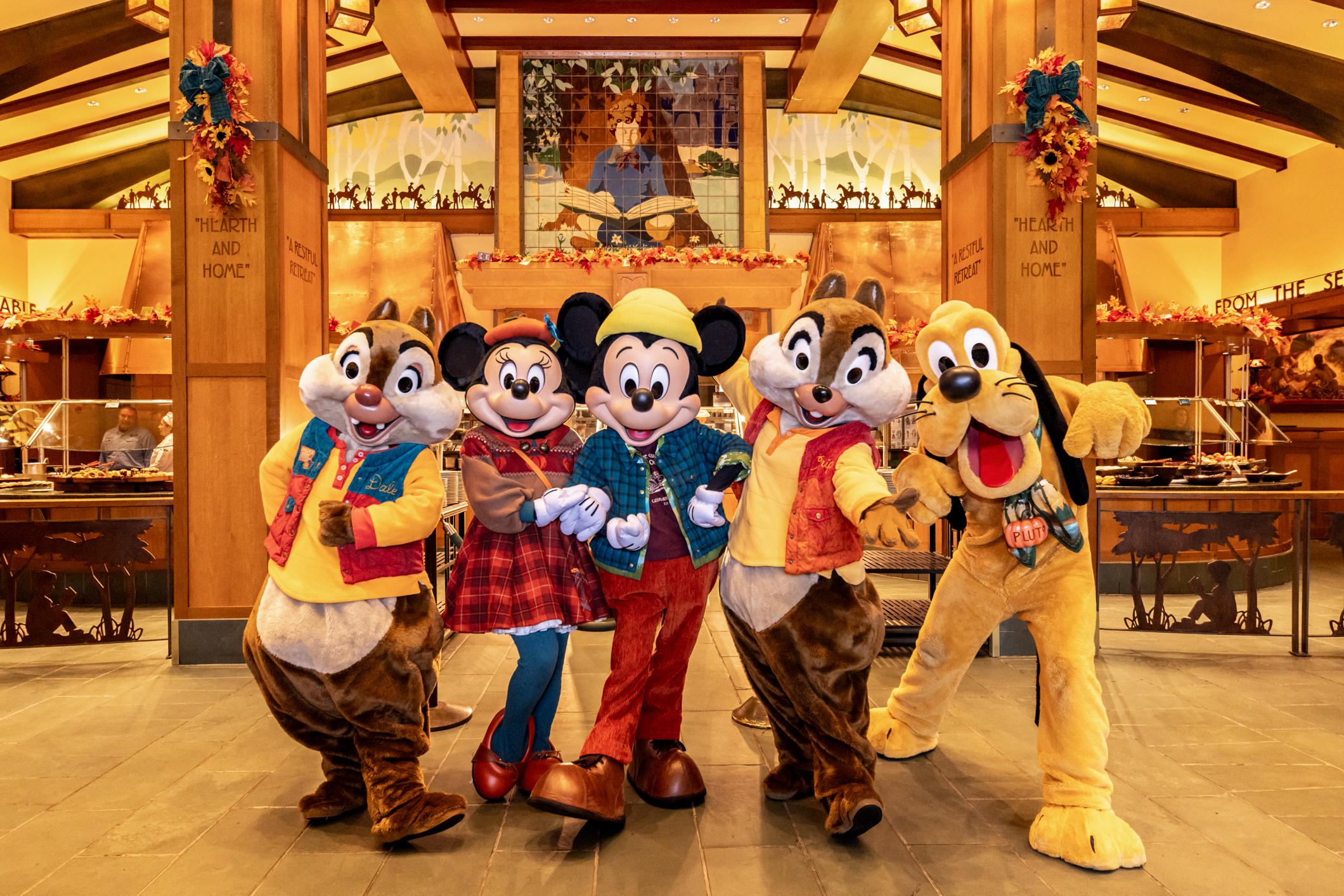 Two chipmunk mascots, Minnie Mouse, Mickey Mouse and Pluto wearing fall outfits pose in a wooden lodge lobby with buffet tables behind them