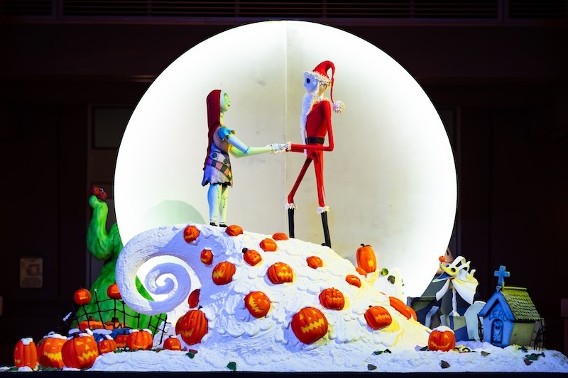 Skeleton in red Santa suit holds hands with a stitched doll atop a curled white hill dotted with jack-o'-lanterns against a glowing circle backdrop