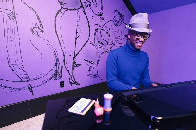 Black man in a gray hat and glasses plays a piano under purple light, sketch mural of bass and drum players on the wall behind him.