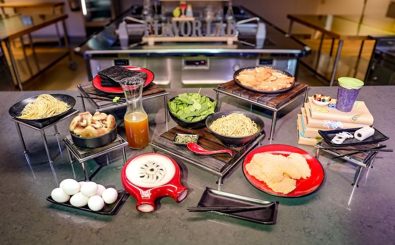 Cooking ingredients on metal stands in a lab kitchen labeled Flavor Lab: noodles, mushrooms, spinach, broth, eggs, breaded cutlets and a smoothie
