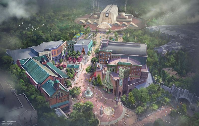 Pedestrian plaza with central fountain, flanked by two-story storefronts and trees, leading to a large industrial-style structure amid forest.
