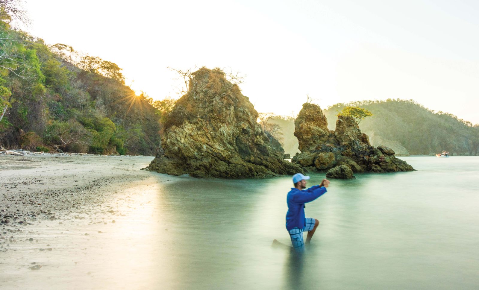 Person wades in shallow water near rocky outcrops on a sandy beach at sunset.