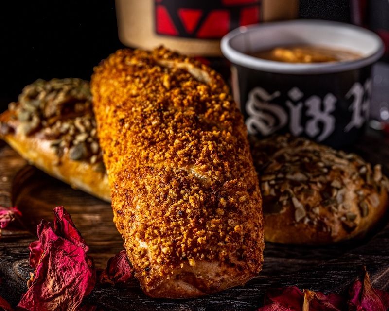 Three assorted bread rolls on a wooden board, including a spice-coated roll, with a cup and dried rose petals nearby