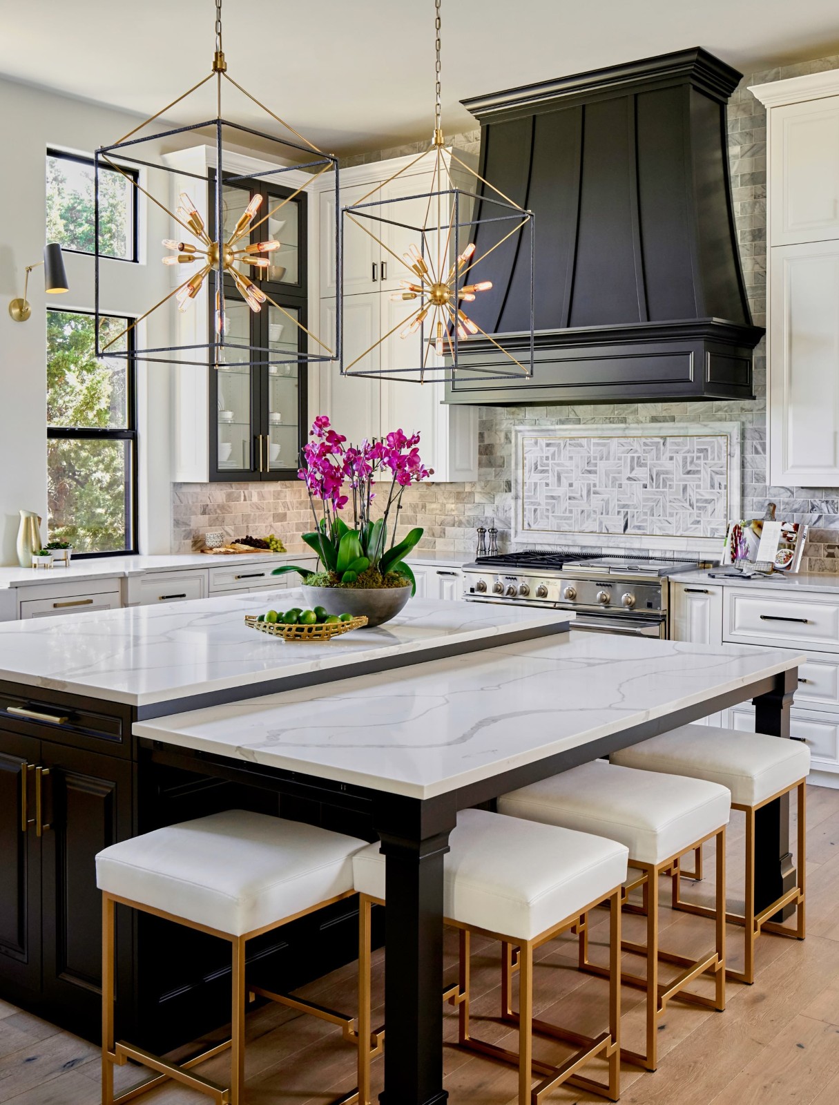Modern kitchen with white marble counters, black hood, gold stools, orchids