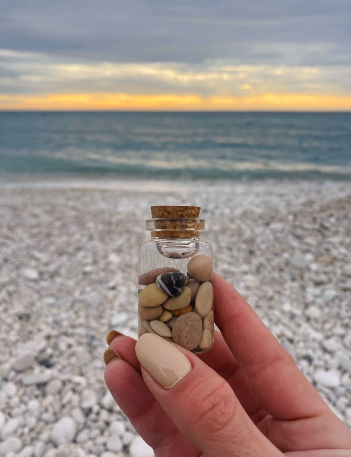 Hand holding a tiny glass bottle filled with beach stones and seawater on the shoreline