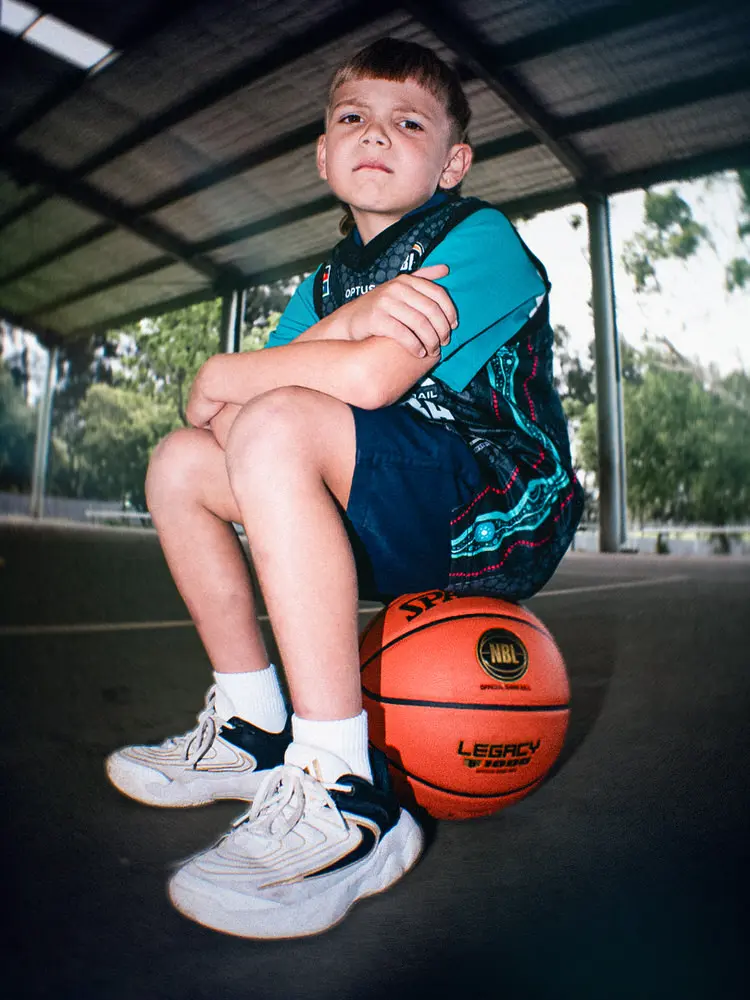 A young participant sits on a basketball, arms folded, wearing the NBL27 Indigenous Jersey.