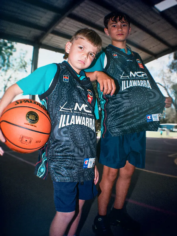 Navi and Tom stand together under a covered court, one holding a basketball, both wearing the NBL27 Indigenous Jersey.