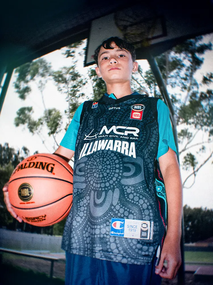 A participant holds a basketball at his side, showing the front of the jersey with MCR and Illawarra across the chest.